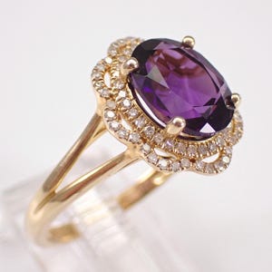 May include: A gold ring with a large, oval purple gemstone and a halo of smaller, clear stones. The band splits into two delicate prongs. The ring's design is classic and elegant. The ring is set against a white background.
