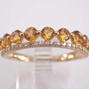 Puede incluir: Un anillo de oro con dos filas de piedras preciosas. La fila superior tiene piedras redondas de color ámbar, mientras que la fila inferior está adornada con piedras más pequeñas y transparentes. El diseño del anillo se destaca sobre un fondo neutro.