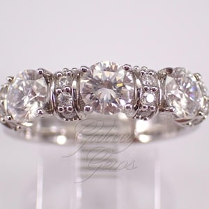 May include: A silver-toned ring featuring three large, round, clear faceted stones set in a row. Smaller, round, clear stones are embedded in the metal band between the larger stones. The text Galaxy Gems is visible in the lower center of the image.