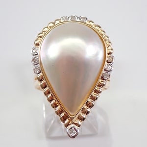 May include: A pear-shaped ring featuring a large, lustrous pearl. The pearl is set in a gold band with a beaded edge and accented with small diamonds. The ring is displayed on a clear stand.