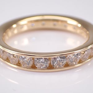 May include: A gold-colored ring with a band of clear, round-cut gemstones. The ring has a smooth, polished finish and a channel setting for the gemstones. The gemstones are evenly spaced around the band.