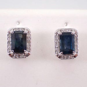 Pode incluir: Um par de brincos de prata com uma pedra preciosa azul escura cercada por pedras preciosas brancas menores.