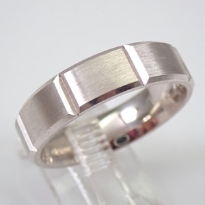 May include: A silver-colored ring with a rectangular design. The ring has a brushed metal finish and a series of rectangular cutouts around the band. The ring is displayed on a clear stand against a white background.