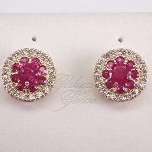 May include: A pair of gold stud earrings. Each earring has a cluster of bright red gemstones at the centre, encircled by a halo of smaller, clear gemstones. The earrings are set against a white background.