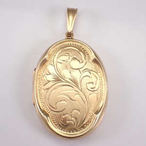 Puede incluir: Un relicario ovalado dorado con un diseño floral ornamentado. El relicario tiene un borde decorativo y un lazo para una cadena. La superficie del relicario está pulida y refleja la luz.