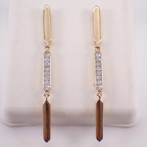 May include: A pair of long drop earrings featuring a vertical design with three connected segments. The top and bottom segments are polished gold, while the center segment is silver-toned and inlaid with a row of small, clear, round-cut stones.