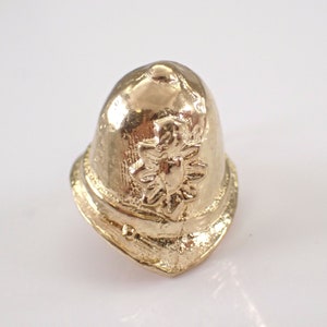 May include: A gold-colored metal replica of a British police officer's helmet. The helmet has a raised design of a crown and a star.