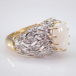 May include: A gold ring with a large oval opal gemstone. The ring has a detailed silver filigree design with small diamonds. The opal is a creamy white colour, and the ring has a classic, elegant style.