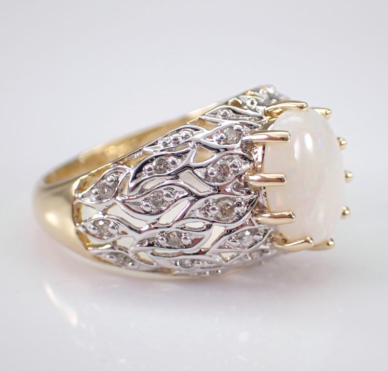 May include: A gold ring with a large oval opal gemstone. The ring has a detailed silver filigree design with small diamonds. The opal is a creamy white colour, and the ring has a classic, elegant style.