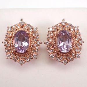 May include: A pair of rose gold earrings. Each earring features an oval, light purple gemstone surrounded by small, clear gemstones. The earrings have a detailed, ornate design, with a halo of smaller stones around the central gem.