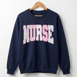 Könnte beinhalten: Marineblaues Sweatshirt mit dem Wort "NURSE" in großen Buchstaben auf der Brust. Die Buchstaben sind aus Patchwork-Stoff mit floralen, Vichy- und gemusterten Designs in Rosa, Weiß und Blau gefertigt. Das Sweatshirt hängt an einem Holzbügel.