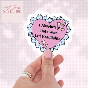 May include: A heart-shaped sticker with the text "I Absolutely Hate Your Led Headlights." The pink heart is outlined with a blue lace design and adorned with yellow stars. The sticker also features the text "Soft Heart Steel Spine."