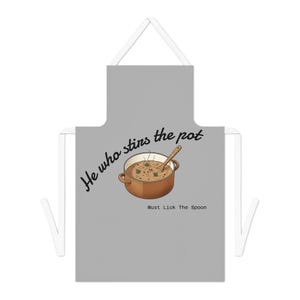 Puede incluir: Delantal gris con correas blancas. Presenta una ilustración de dibujos animados de una olla de sopa con una cuchara de madera. El texto dice "He who stirs the pot" y "Must Lick The Spoon".