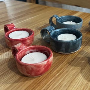 Puede incluir: Una colección de portavelas de cerámica en varios colores, incluyendo rojo, azul y marrón. Cada portavelas tiene un asa pequeña y contiene una vela de té blanca. Los portavelas están colocados sobre una superficie de madera.