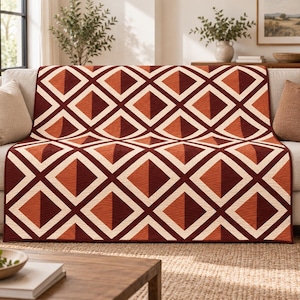 Op de afbeelding: Een rechthoekige quilt met een herhalend geometrisch patroon van ruiten in tinten diep bordeaux, gebrand oranje en crème. Het ontwerp creëert een driedimensionaal effect met kruisende lijnen en contrasterende kleurblokken over het geheel.