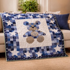 May include: A blue and white patchwork quilt with a teddy bear design. The quilt has a white centre with a bear, surrounded by a border of blue squares and white stars. The quilt is draped over a beige sofa.