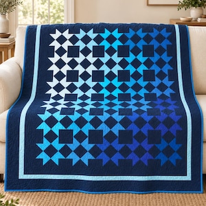 Puede incluir: Una colcha rectangular con un patrón geométrico de estrellas y cuadrados en un degradado de tonos azules. El diseño transita de azul claro a azul marino profundo, enmarcado por un borde azul marino sólido y un borde interior azul claro.