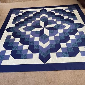 Puede incluir: Un edredón grande y cuadrado con un diseño geométrico en tonos azules y blancos. El edredón presenta un patrón central de estrella y un borde azul oscuro. El diseño incorpora cuadrados, triángulos y formas de chevrón.