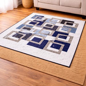 May include: A geometric quilt with a white background and a pattern of interlocking squares in shades of blue, gray, and black. The quilt is placed on a woven rug and hardwood floor, creating a cozy home decor scene.