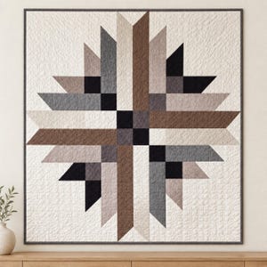 May include: A square quilt featuring a central starburst pattern composed of geometric shapes in shades of black, charcoal gray, brown, and beige. The quilt has a textured, quilted surface and is framed by a thin, dark gray border.