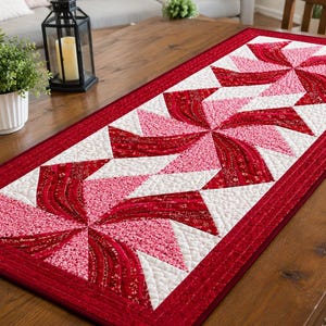 May include: A rectangular quilted table runner featuring a repeating pinwheel pattern in shades of red, pink, and white. The fabric displays intricate stitching details and a solid red border, creating a structured design across the entire surface.