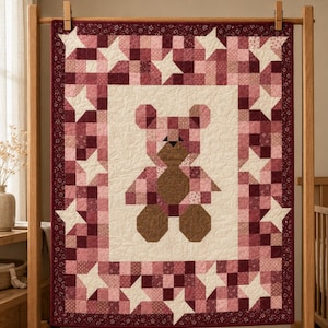 May include: A rectangular quilt featuring a central teddy bear motif pieced together from various shades of pink and brown fabric. The bear is set against a cream background, surrounded by a border of pink and burgundy patchwork star patterns.