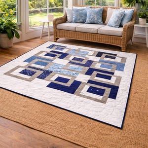 May include: A geometric quilt with a white background and a pattern of blue and gray squares and rectangles. The quilt is placed on a natural fiber rug, with a wicker sofa and blue pillows in the background. The room has large windows.
