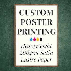 May include: A rectangular poster in a brown wooden frame with white background and dark gray text reading CUSTOM POSTER PRINTING, followed by four colored ink drops in cyan, magenta, yellow, and black, and text Heavyweight 260gsm Satin Lustre Paper.