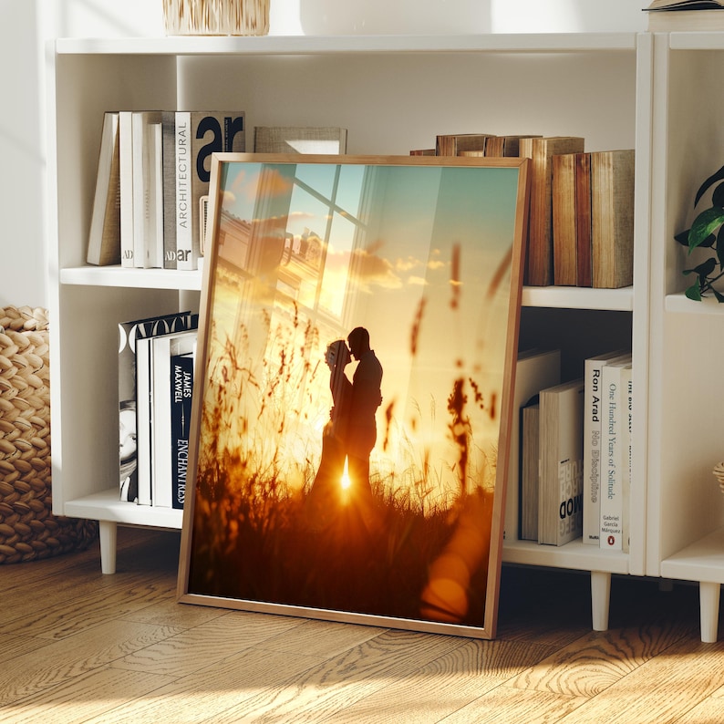 May include: A rectangular framed print featuring a silhouette of two people embracing in a field of tall grass during a golden sunset. The warm orange and yellow light creates a glowing effect around the figures, with tall stalks of grass visible in the foreground and background.