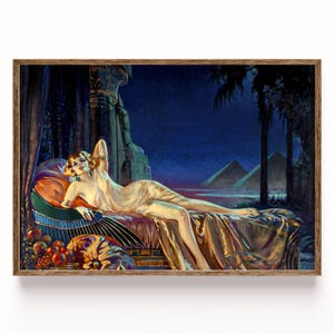 May include: Vintage art print in a wooden frame. The artwork depicts a woman in a flowing gown reclining on a bed with gold linens. The background features pyramids, palm trees, and a dark blue sky, creating an exotic, historical scene.
