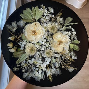 May include: A round, black decorative tray featuring a floral arrangement. The arrangement includes cream-colored roses, white blossoms, and green foliage. The tray's glossy surface enhances the colors of the dried flowers.