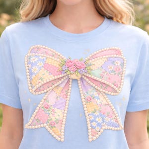 May include: A pale blue t-shirt with a large bow design. The bow is made of patchwork fabric in pastel shades, edged with a pearl trim. The bow is centered on the shirt, with the text "HIGH QUALITY 300 DPI" and "PNG+PDF" at the top and bottom.