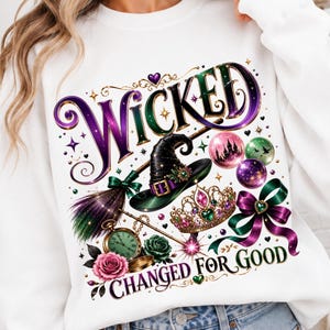 May include: A white sweatshirt featuring a graphic print with the word WICKED in purple and green lettering. The design includes a black witch hat, a broom, a gold crown with jewels, a pocket watch, roses, and the text CHANGED FOR GOOD.