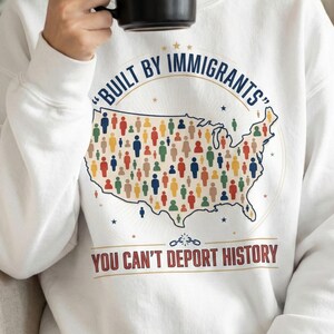 May include: White sweatshirt with the text "BUILT BY IMMIGRANTS" in blue, and "YOU CAN'T DEPORT HISTORY" in brown. The design includes a map of the United States filled with colorful figures.