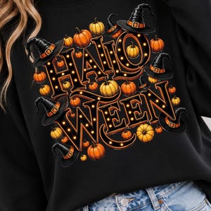 May include: A black sweatshirt featuring a graphic print of the word HALLO WEEN in large, bold, orange-outlined letters with small light bulbs inside. The design is surrounded by numerous orange pumpkins and several black witch hats.