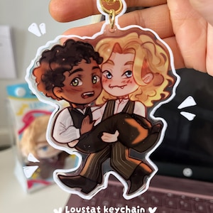 Op de afbeelding: Een sleutelhanger met een cartoonillustratie van twee personages, waarvan de ene de andere draagt. De personages hebben donkere en lichte huidtinten, de ene met krullend donker haar en de andere met lang blond haar. De sleutelhanger heeft een goudkleurige sluiting en de tekst "Loustast keychain".