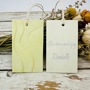 May include: Two rectangular, light green tags with cream-colored rope loops. One tag has a wavy pattern, the other has the text "Slow down and enjoy Danielle" in a script font. Dried flowers are in the background.