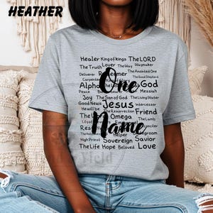 Peut inclure: T-shirt gris chiné avec les mots "One Name" en écriture noire. Le t-shirt présente une liste de mots tels que "Jesus", "God", "Friend" et "Savior" en noir.