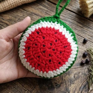 May include: A handmade crochet watermelon-shaped item with a green border, white middle, and red center with black seed details. The item has a green loop for hanging. The item is held in a hand.