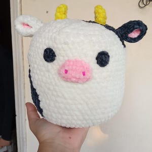 May include: A handmade, plush cow toy with a white body, black spots, and pink accents. The cow has black eyes, a pink nose, yellow horns, and black and pink ears. The toy is held in a person's hand.