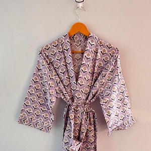 May include: A lilac-colored robe with a floral pattern, hanging on a wooden hanger. The robe features a repeating design of stylized flowers in white and brown. The robe is tied at the waist with a matching fabric belt.