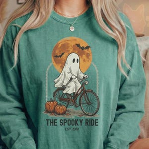 Könnte beinhalten: Ein hellgrünes Langarmshirt mit einer Grafik, die ein Gespenst auf einem Fahrrad vor einem großen orangefarbenen Mond zeigt. Zwei Kürbisse liegen auf dem Boden und Fledermäuse fliegen darüber. Der Text lautet THE SPOOKY RIDE EST. 1931.