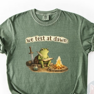 May include: Moss green t-shirt featuring a whimsical illustration of a frog writing by a campfire with a sword and backpack. The text "we test at dawn" is above the frog. The shirt has a classic crew neck and short sleeves.