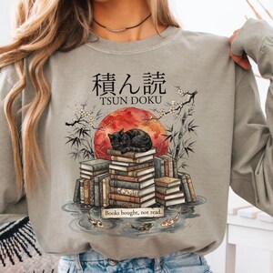 Op de afbeelding: Een zandsteenkleurig sweatshirt met lange mouwen en een grafische print van een zwarte kat die op een stapel boeken slaapt. Het ontwerp bevat een rode zon, bamboetakken, koikarpers in water en de tekst "積ん読 TSUN DOKU" en "Books bought, not read."
