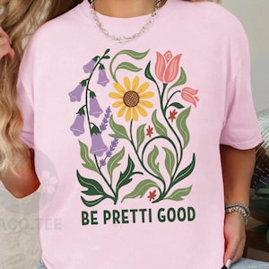 Può includere: T-shirt rosa chiaro con un design floreale con un girasole giallo, tulipani rosa e fiori viola a forma di campana. Il testo "BE PRETTI GOOD" è stampato sotto. La maglietta è ampia.