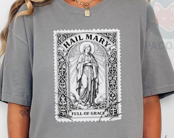 May include: A gray t-shirt featuring a black and white graphic of a religious image with the text "HAIL MARY" and "FULL OF GRACE". The shirt is a casual style.