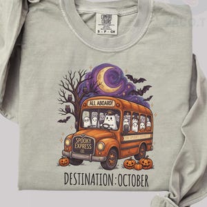 May include: A light gray long-sleeve crewneck sweatshirt featuring a graphic of an orange bus labeled Spooky Express Co. filled with ghosts. The design includes a crescent moon, bats, a bare tree, and pumpkins. Text reads All Aboard and Destination: October.
