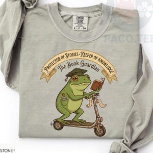 Op de afbeelding: Een zandkleurig t-shirt met lange mouwen en een afbeelding van een groene kikker met een afstudeerhoed op een step, die een boek vasthoudt. De tekst luidt Protector of Stories, Keeper of Knowledge en The Book Guardian in bruin.