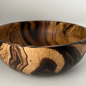 May include: A wooden bowl featuring a striking grain pattern in brown and black hues. The bowl has a smooth, rounded form and a polished finish, ideal for serving food or as a decorative item.