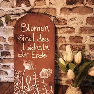 May include: A brown wooden sign with the text "Blumen sind das Lächeln der Erde" in white script, along with white floral illustrations. A vase of white tulips sits to the right of the sign.